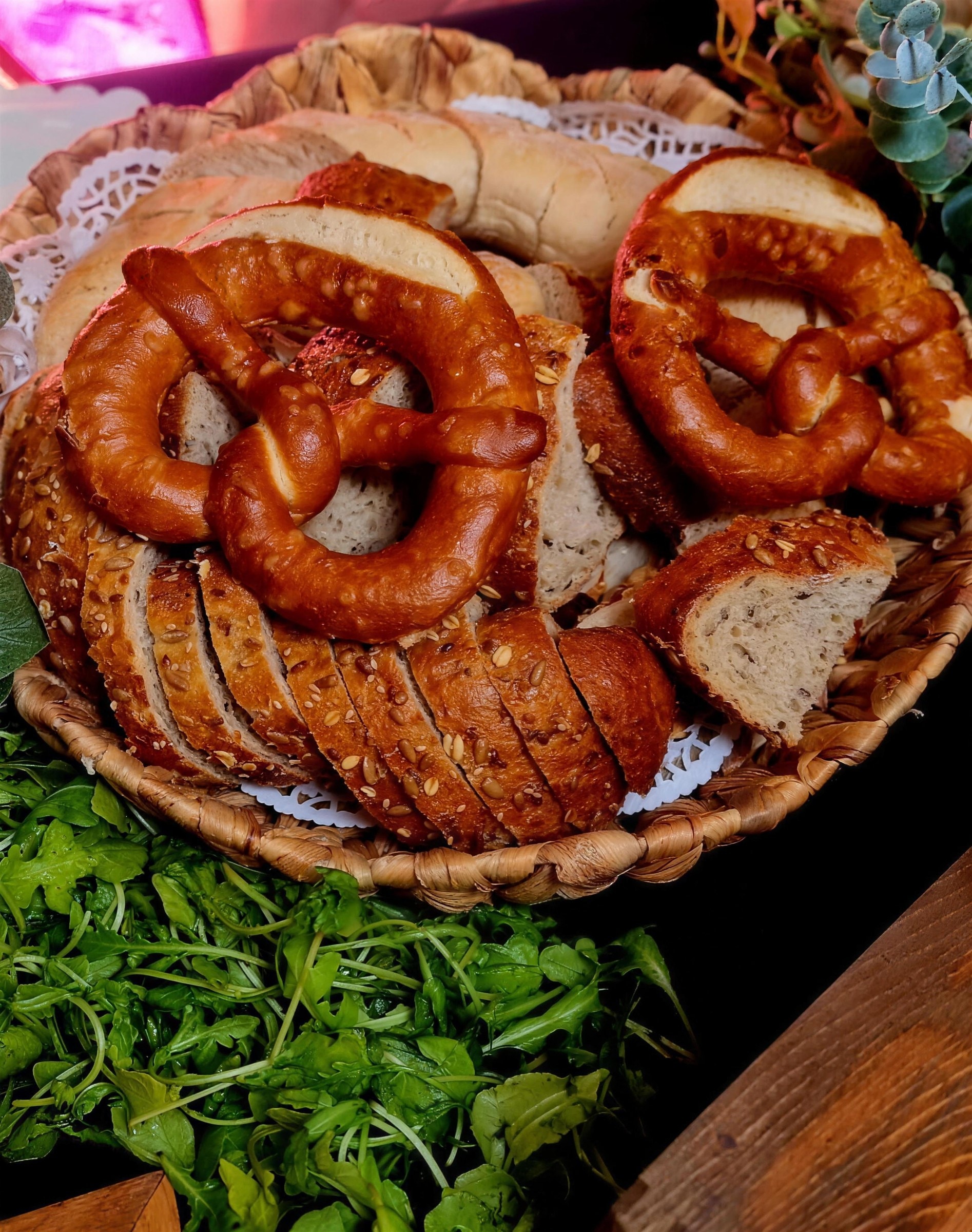 Basket of sliced seeded bread and soft pretzels resting on a bed of fresh rocket